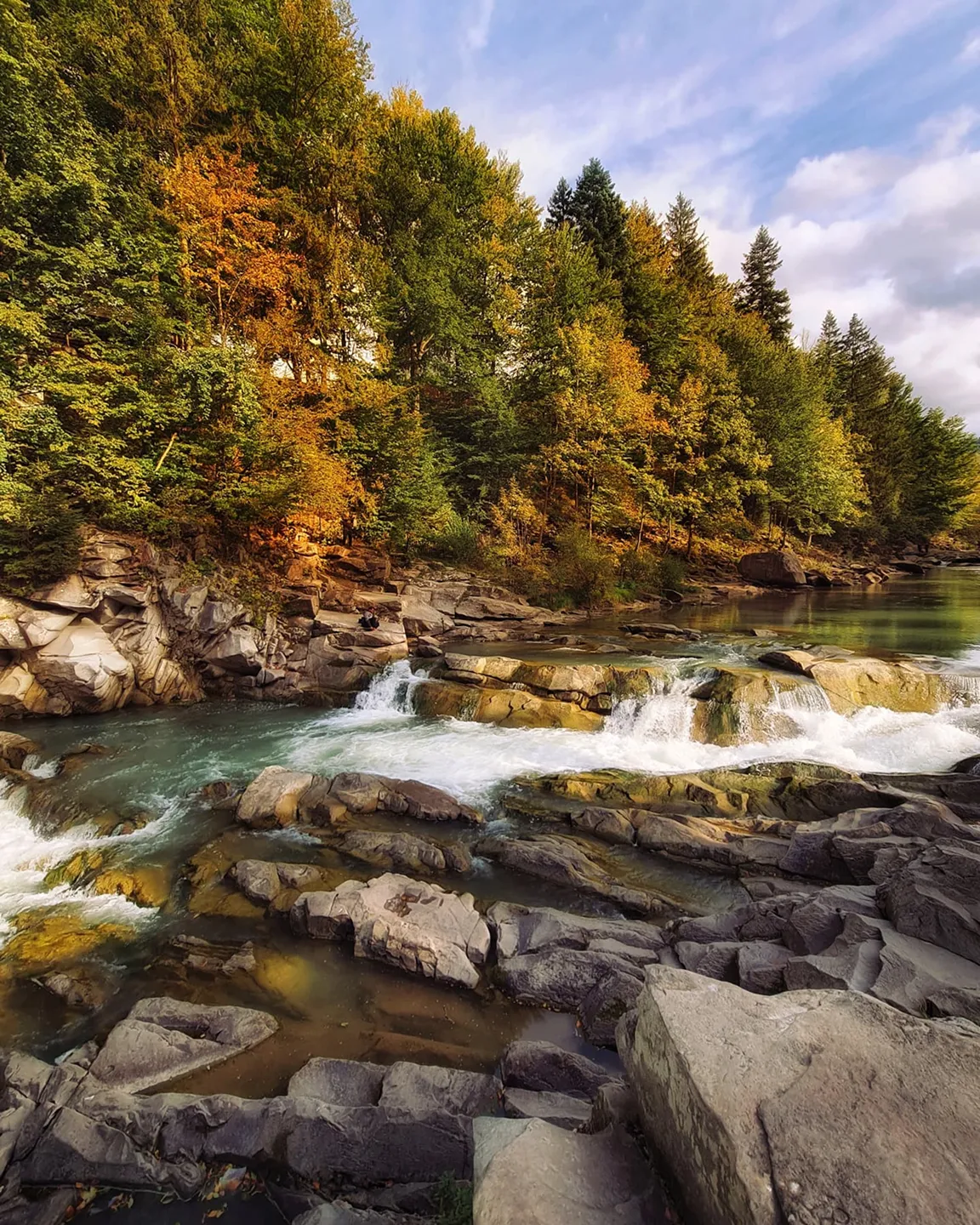 Une rivière à seuils entourée d'une forêt d'automne aux feuilles jaunes et vertes, sous un ciel bleu parsemé de nuages.